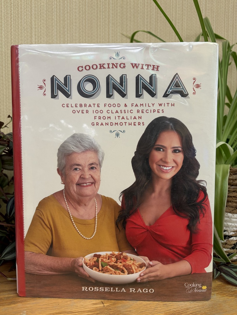 Cooking with Nonna cookbook on display on a shelf with plants behind it.